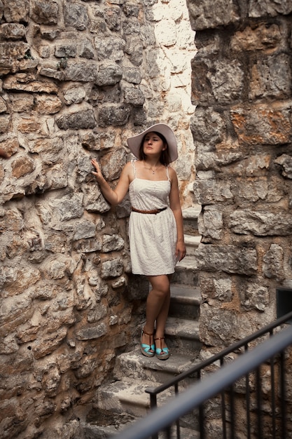 Bella giovane donna in vestito e cappello che cammina all'antico castello di pietra