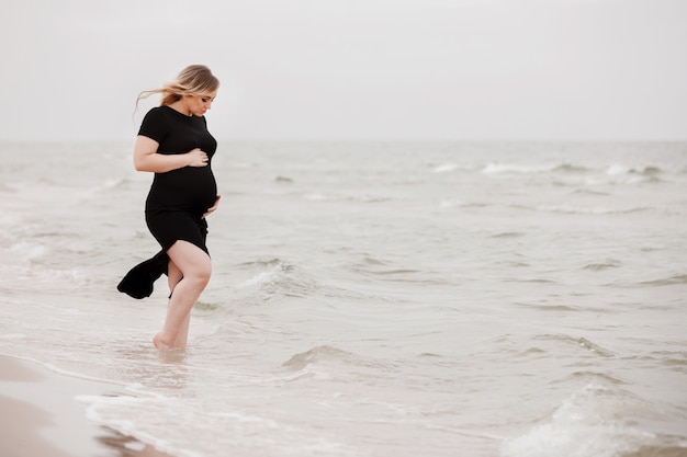 Bella giovane donna incinta che porta vestito nero che cammina sulla spiaggia