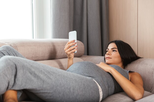 Bella giovane donna incinta posa su un divano a casa, utilizzando il telefono cellulare