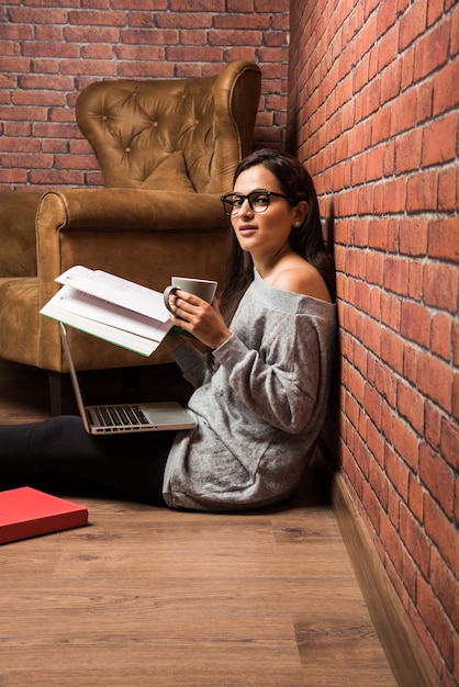 Bella giovane donna indiana o asiatica che studia con un libro e un laptop sul pavimento