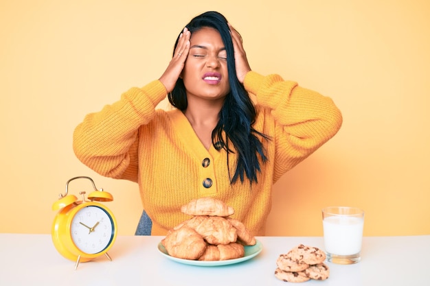 Bella giovane donna latina con i capelli lunghi seduta sul tavolo a fare colazione che soffre di mal di testa disperata e stressata perché dolore ed emicrania. mani sulla testa.