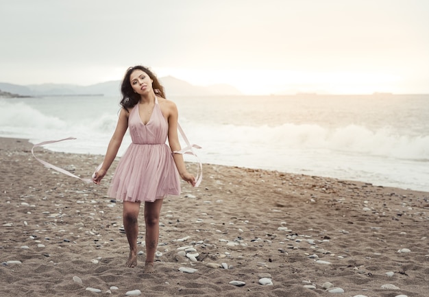 Bella giovane donna latina con lunghi capelli neri, che corre lungo la spiaggia al tramonto, indossa un elegante abito rosa, gioca e balla con i nastri in mano