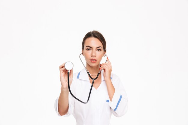 Bella giovane donna medico indossa uniforme in piedi isolato su muro bianco, utilizzando uno stetoscopio