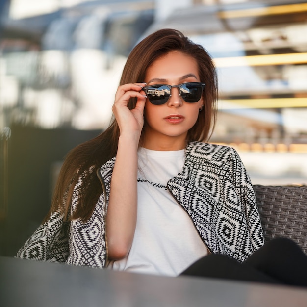 Bella giovane donna modello con occhiali da sole in una maglietta bianca con un bomber di moda si siede in un caffè sulla strada