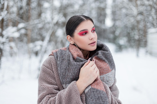 Bella giovane donna modello con trucco luminoso colorato e ombretti rosa in abiti invernali alla moda con pelliccia e sciarpa ha sullo sfondo di una foresta di neve