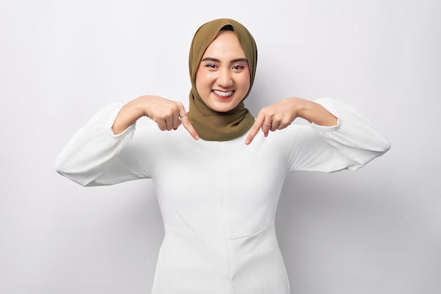 Bella giovane donna musulmana asiatica araba sorridente che indossa l'hijab in piedi puntando il dito verso il basso guardando la telecamera isolata su sfondo bianco concetto di stile di vita religioso persone