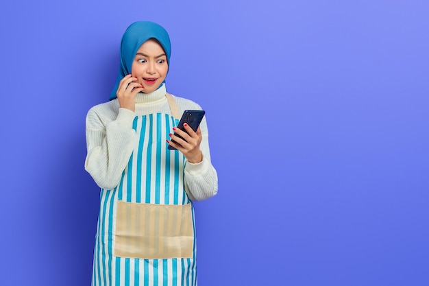 Bella giovane donna musulmana asiatica che indossa hijab e grembiule, guardando ig promo su smartphone con espressione sorpresa isolata su sfondo viola. Concetto di stile di vita musulmano casalinga della gente