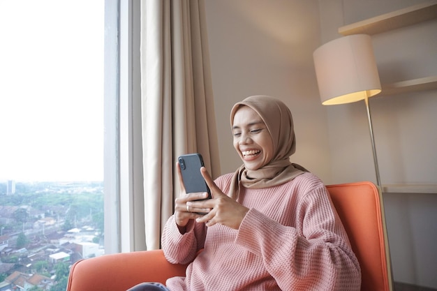 bella giovane donna musulmana asiatica che indossa l'hijab mentre utilizza lo smartphone seduto vicino alla finestra