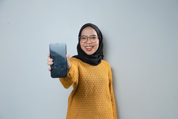 Bella giovane donna musulmana asiatica che indossa maglione arancione e occhiali che puntano il telefono alla macchina fotografica