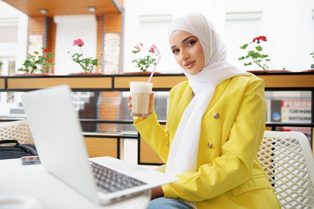 Bella giovane donna musulmana che usa il computer portatile mentre è seduta al bar