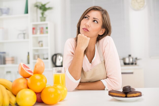 Bella giovane donna nella decisione della cucina tra frutta o torta al cioccolato dolce.