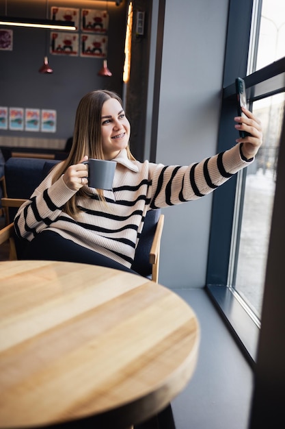 Bella giovane donna positiva in abiti casual che sorride, beve una tazza di caffè e tiene un telefono in mano mentre trascorre del tempo nella caffetteria