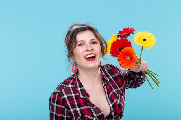 Bella giovane donna positiva in una camicia a quadri e una benda che fiuta bellissimi fiori luminosi di gerbera in posa su una superficie blu