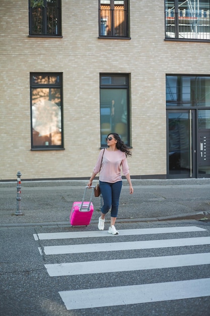 Bella giovane donna riccia con una piccola valigia rosa ha fretta di andare in vacanza Una modella con i capelli lunghi attraversa la strada in una città europea Viaggi