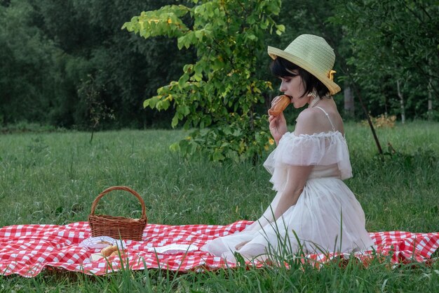 Bella giovane donna romantica con i capelli corti scuri in un cappello di paglia e vestito bianco è seduta su un picnic nella natura