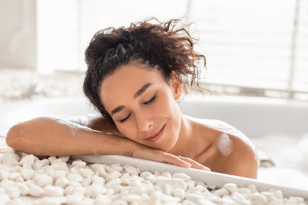 Bella giovane donna sdraiata nel bagno di bolle con gli occhi chiusi a casa concetto di bellezza e benessere