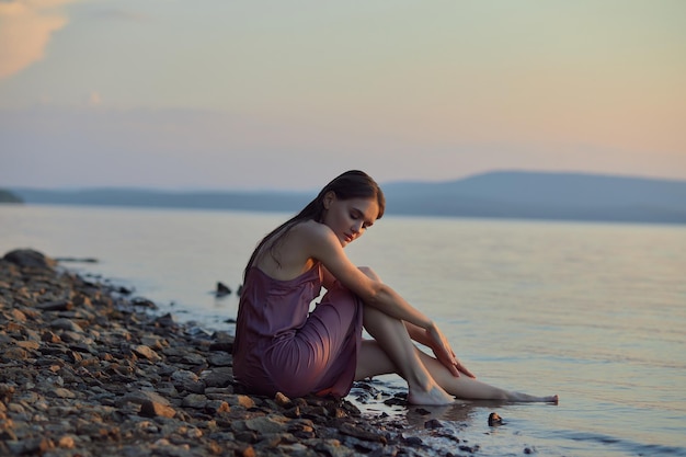 Bella giovane donna seduta sulla riva del lago in abito estivo al tramonto Ritratto di una romantica ragazza bagnata al tramonto caldo sole bellezza naturale di una donna