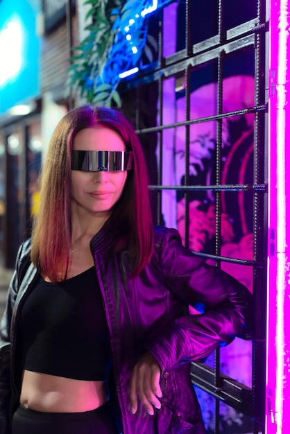Bella giovane donna sfocata in occhiali cosmici in piedi accanto alla luce al neon colorata