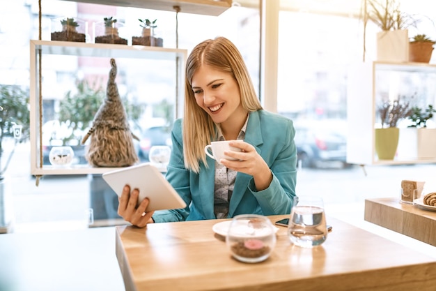 Bella giovane donna sorridente che beve caffè in un caffè. Sta navigando in Internet su una tavoletta digitale.