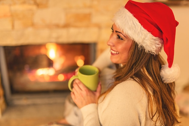 Bella giovane donna sorridente che gode di una tazza di tè davanti al camino nella sera di Natale.