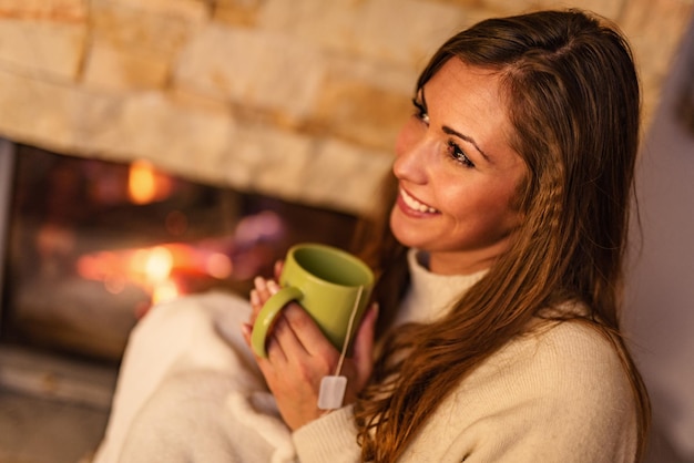 Bella giovane donna sorridente che gode di una tazza di tè davanti al camino.