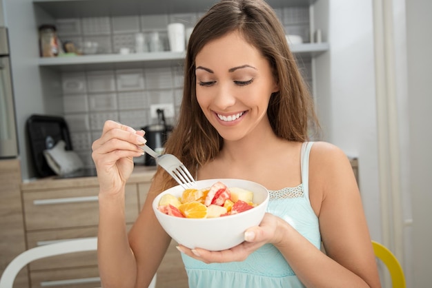 Bella giovane donna sorridente che mangia insalata di frutta in cucina.