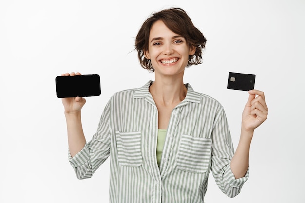 Bella giovane donna sorridente, che mostra lo schermo orizzontale dello smartphone e la carta di credito, l'interfaccia dell'app, l'app di finanziamento dei pagamenti, in piedi su sfondo bianco.