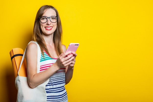 Bella giovane donna sorridente con karimat in una borsa che tiene un telefono su uno sfondo giallo.