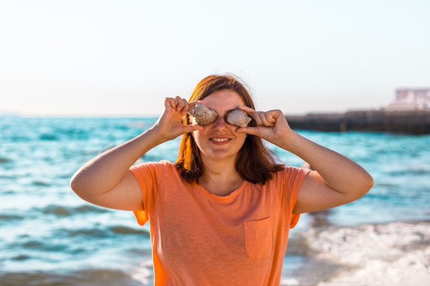 Bella giovane donna sorridente e tenendo due conchiglie sul viso con gli occhi sulla spiaggia