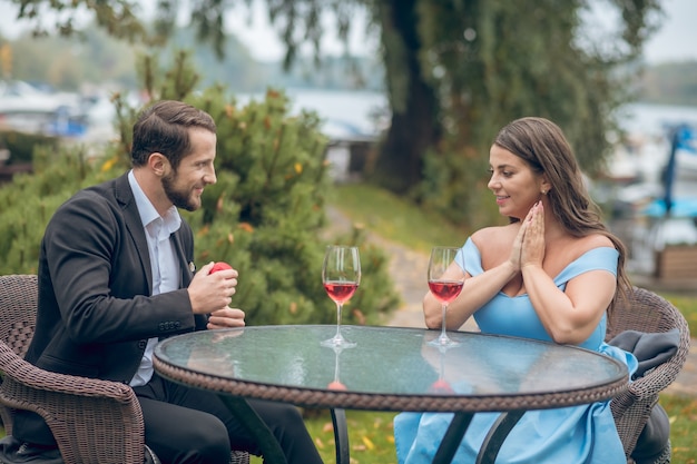 Bella giovane donna sorridente e uomo con regalo seduto al tavolo uno di fronte all'altro con bicchiere di vino all'aperto