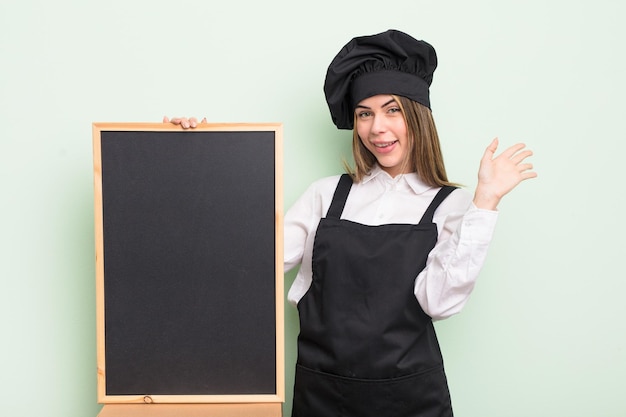 bella giovane donna sorridente felice, agitando la mano, dandoti il benvenuto e salutandoti. chef con lavagna