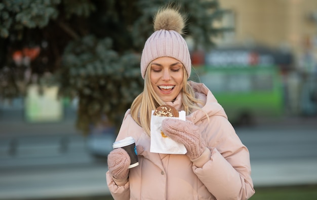 Bella giovane donna sorridente in abiti caldi con una tazza di tè o caffè caldo pugno. Donna che mangia i biscotti all'aperto.