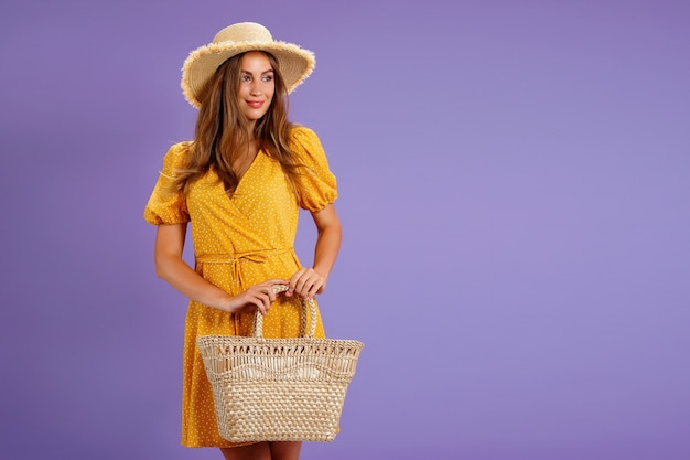 Bella giovane donna sorridente in borsa gialla del cappello di paglia di estate del vestito isolata su fondo viola pastello