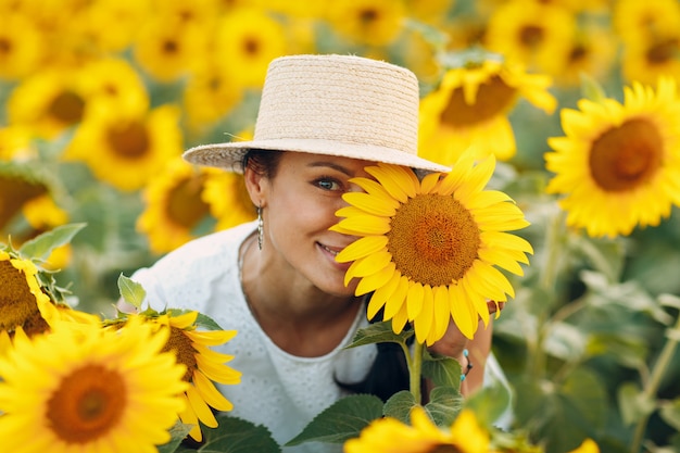 Bella giovane donna sorridente in un cappello con un fiore sugli occhi e sul viso su un campo di girasoli