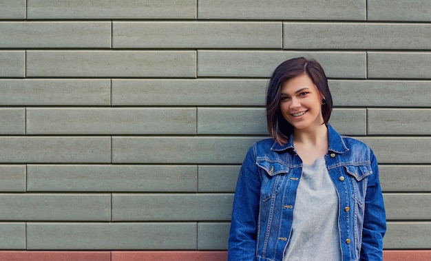 Bella giovane donna sorridente in una giacca di jeans blu vicino a un muro di mattoni con texture.