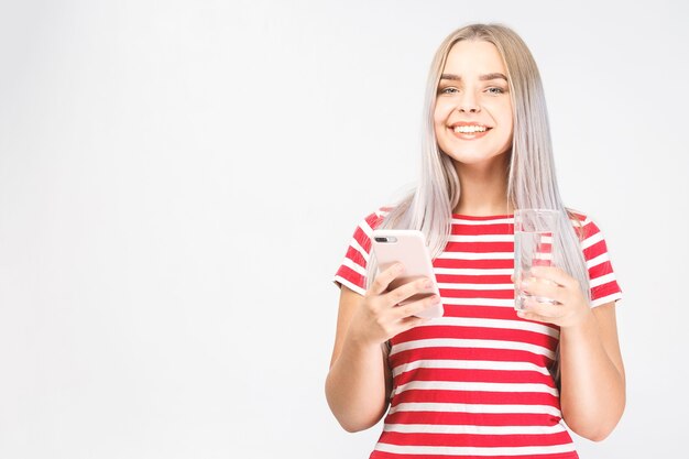 Bella giovane donna sorridente si trova su sfondo bianco e guarda al telefono con una tazza di acqua fredda con il telefono cellulare. Isolato, mandare SMS.