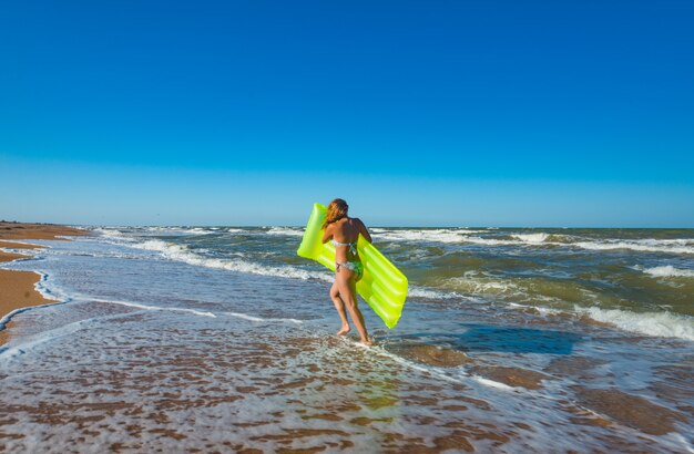 Bella giovane donna sottile in costume da bagno corre lungo la spiaggia con un materasso ad aria