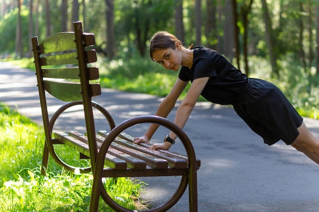 Bella giovane donna sportiva in maglietta nera, pantaloncini neri e scarpe da ginnastica rosa che si riscaldano esercitando i tricipiti e il torace facendo flessioni dalla panchina tra gli alberi prima di correre