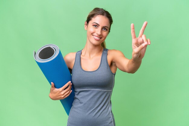 Bella giovane donna sportiva uruguaiana che va alle lezioni di yoga tenendo un tappetino su sfondo isolato sorridendo e mostrando il segno della vittoria