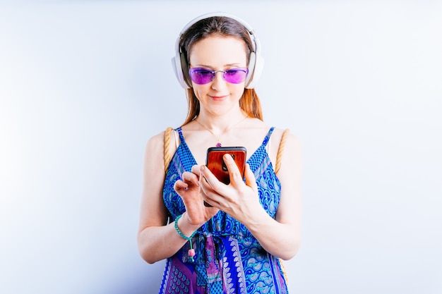 Bella giovane donna su sfondo bianco che indossa occhiali estivi e cuffie wireless guardando smartphone. il suo concetto va in vacanza. offerta. pubblicità con copia spazio