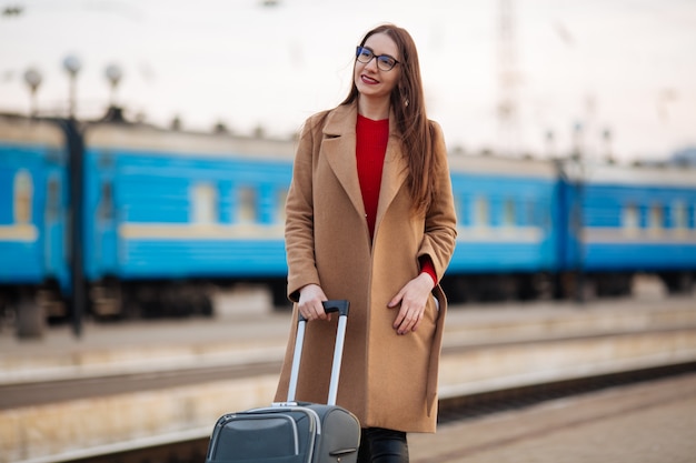 Bella giovane donna turistica con il treno aspettante della valigia alla stazione ferroviaria. Viaggi, viaggi, vacanze, turismo, vacanze