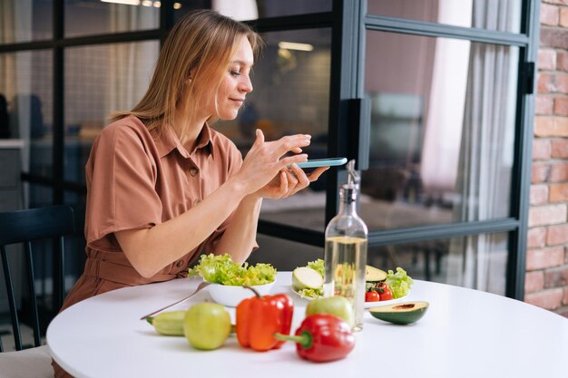 Bella giovane donna utilizza lo smartphone per scattare foto di verdure sdraiate sul tavolo da cucina Concetto di social network Concetto di mangiare sano