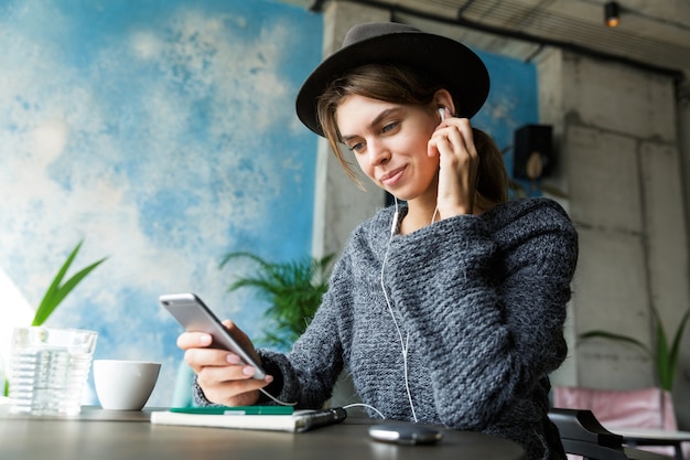Bella giovane donna vestita di maglione e cappello seduto in poltrona al tavolino del bar, ascoltando musica con gli auricolari, utilizzando il telefono cellulare, interni eleganti