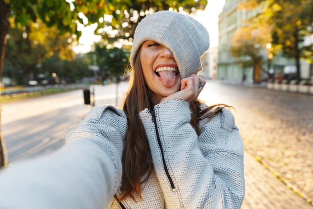 Bella giovane donna vestita in cappotto di autunno e cappello che cammina all'aperto, prendendo un selfie