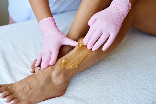 Bella giovane estetista applica pasta di zucchero alla gamba del cliente. Depilazione con shugaring. Estetista che depila giovane donna.