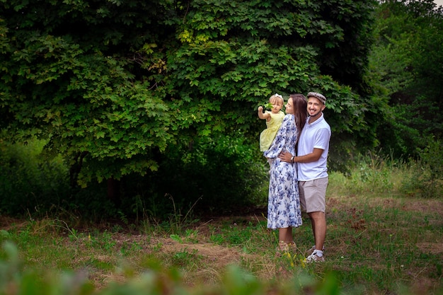 Bella giovane famiglia che porta piccola figlia e si sorride a vicenda nel parco verde durante la calda giornata estiva