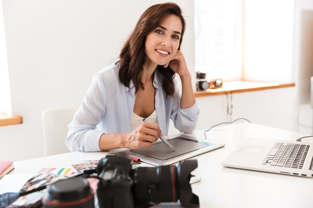 Bella giovane fotografa che lavora nel suo ufficio mentre è seduta alla scrivania, lavorando con il tablet per disegnare