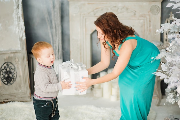 Bella giovane madre castana in vestito blu elegante e bambino sorridente con il regalo di Natale avvolto. intorno albero di natale bianco e regali. Foto glamour del nuovo anno