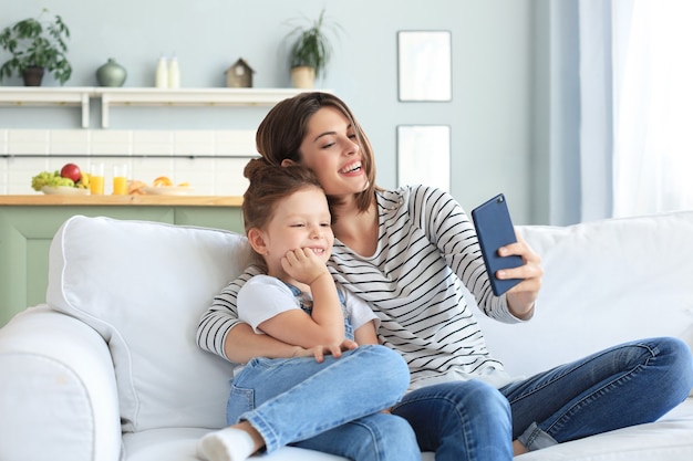 Bella giovane madre e la sua piccola figlia che si fanno un selfie con uno smartphone a casa sul divano.
