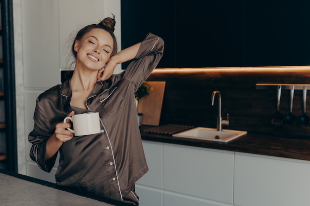 Bella giovane modella femminile rilassata in pigiama accogliente con una tazza di caffè in cucina